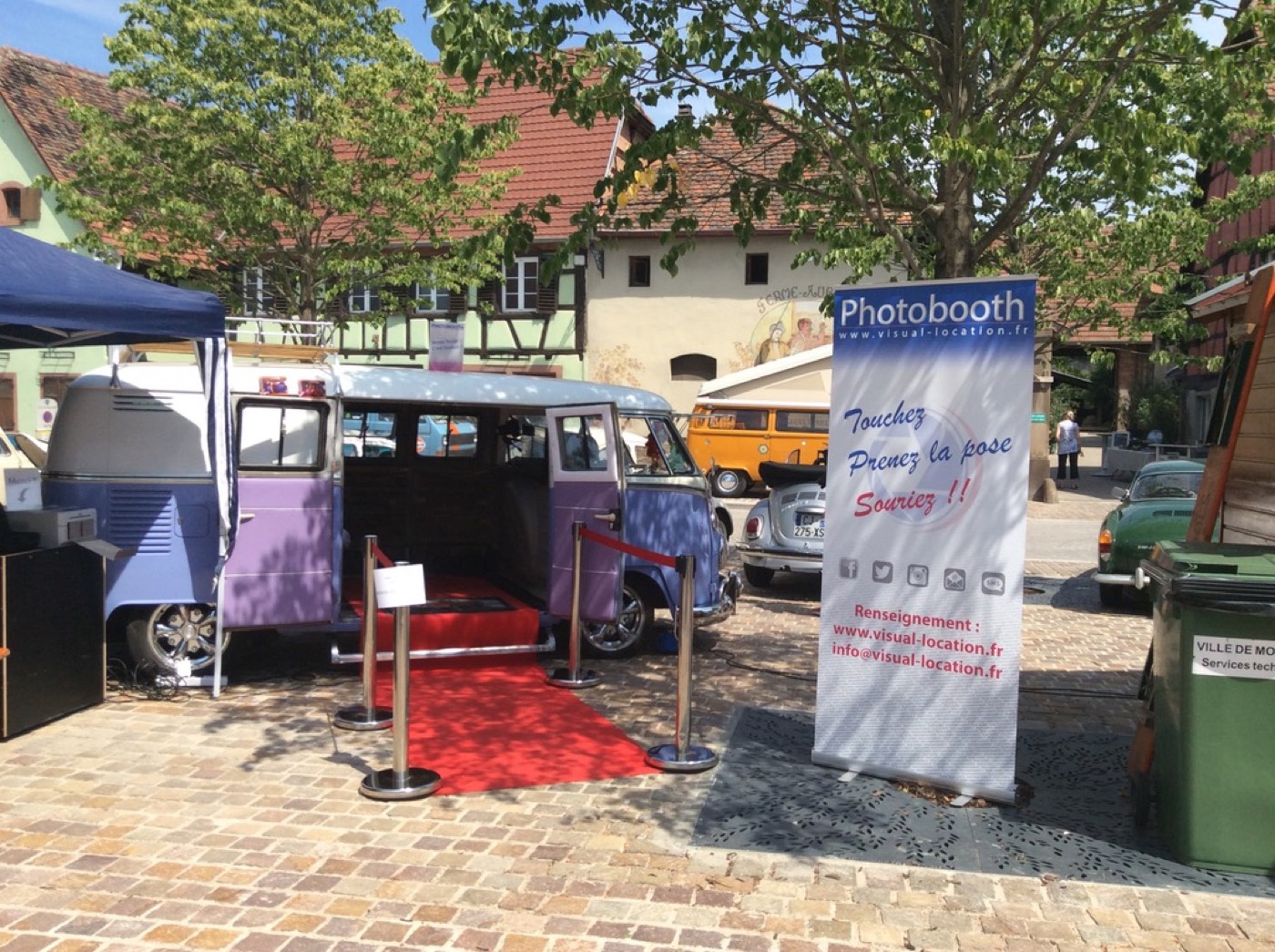 Le combi Volkswagen photobooth installé sur une place de village, tapis rouge et signalétique